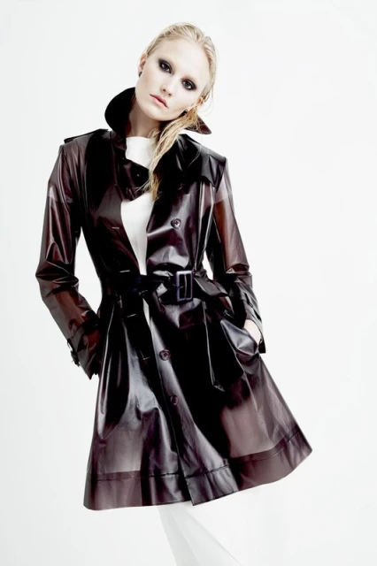 Fashionable woman in a glossy brown trench coat posing in a bright white studio setting, showcasing modern style and clothing design.