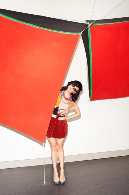Woman in a black top stands against a gray studio backdrop with soft lighting, showcasing fashion and beauty elements.
