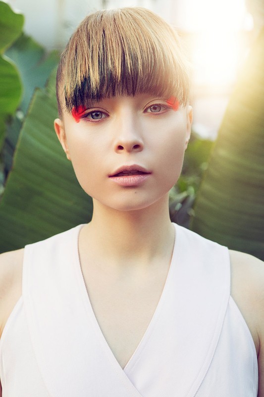 Close-up portrait of a woman with creative red eye makeup, bold blonde bangs, and a light complexion posing against a lush green backdrop.