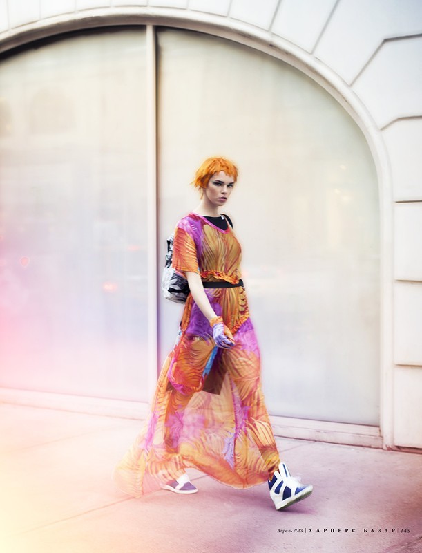 Fashion editorial shot of a young adult woman walking outside wearing a long orange and purple patterned dress and a backpack.