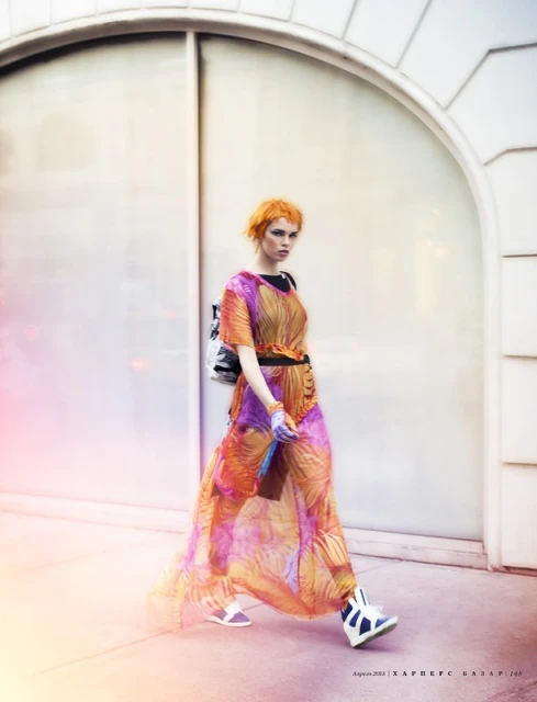 Fashion editorial shot of a young adult woman walking outside wearing a long orange and purple patterned dress and a backpack.
