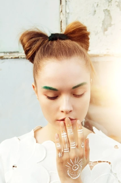 Young woman with creative hair and makeup poses with hands on her face in front of a distressed wall, capturing a fusion of art and beauty.