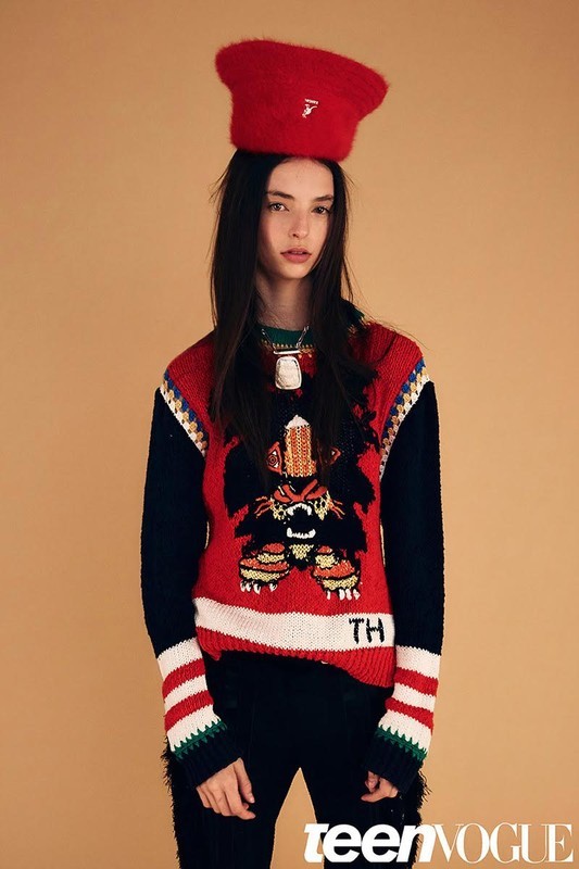 A young woman poses in a red hat and colorful sweater against a neutral background for a fashion editorial, looking directly at the camera.