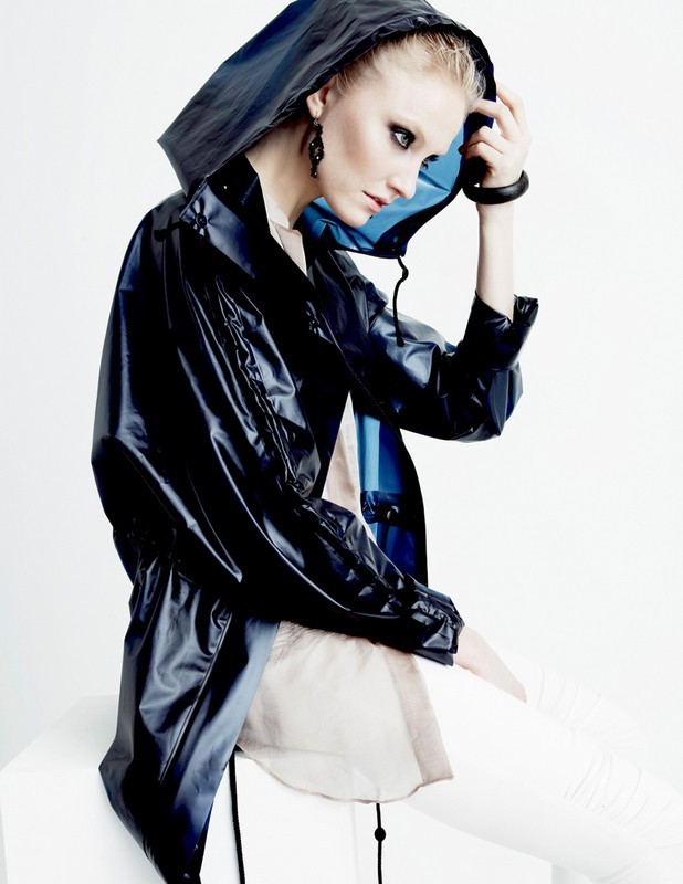 A woman in a hooded, blue-black, shiny jacket with white pants poses in a studio setting against a bright background. Fashion shot.