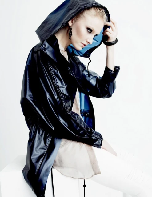 A woman in a hooded, blue-black, shiny jacket with white pants poses in a studio setting against a bright background. Fashion shot.