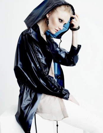 A woman in a hooded, blue-black, shiny jacket with white pants poses in a studio setting against a bright background. Fashion shot.