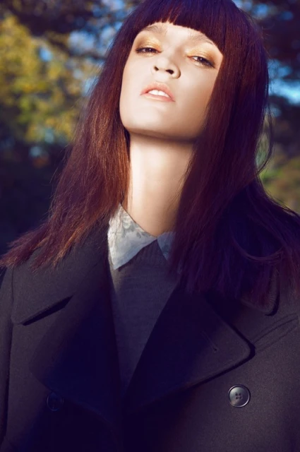 A woman with bangs and a dark coat poses outdoors in a fashion editorial portrait with neutral colors and soft, diffused sunlight.