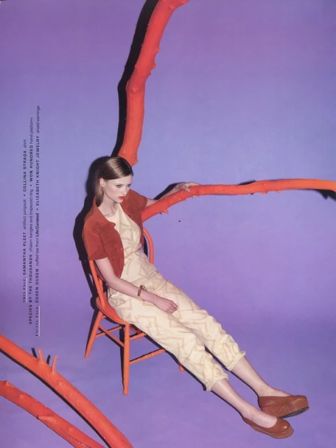 A young woman models a patterned cream jumpsuit with a rust-colored jacket and flats, seated on a matching chair against a purple backdrop.