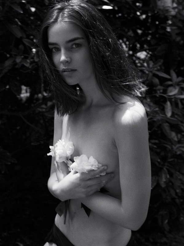 Black and white portrait of a fair-skinned young woman holding white flowers, set against a backdrop of dark green foliage outdoors.