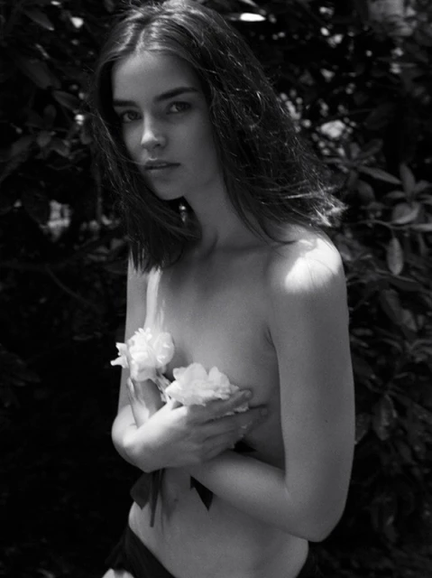 Black and white portrait of a fair-skinned young woman holding white flowers, set against a backdrop of dark green foliage outdoors.