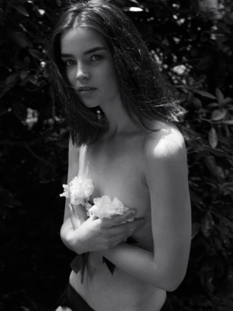Black and white portrait of a fair-skinned young woman holding white flowers, set against a backdrop of dark green foliage outdoors.