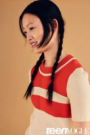 Portrait of a woman with braided dark hair wearing a red and cream striped top against a neutral tan background.