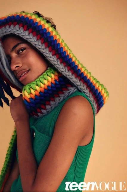 Teen Vogue portrait of a young adult in a green top and a large colorful knitted hood with rainbow accents, set against a neutral backdrop.