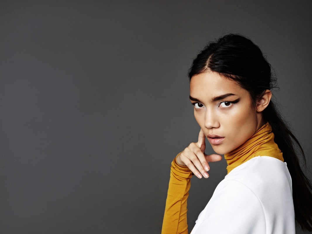 Portrait of a striking young adult woman with dark hair and stylish eye makeup. She's wearing a mustard-yellow and white top.