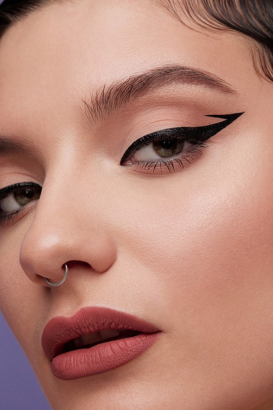 Close-up portrait of a woman with black eyeliner and mauve lipstick against a lilac background, emphasizing makeup and facial features.