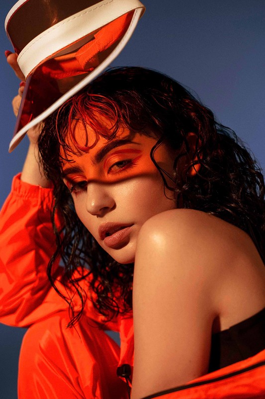 Close-up of a brunette woman with orange visor, wet hair, and orange makeup, posing indoors against a gradient blue background.