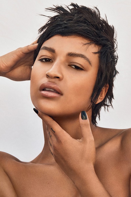 Close-up portrait of a person with short dark hair and light skin, hands near the neck, set against a pale background.