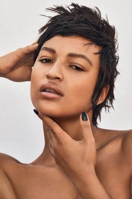 Close-up portrait of a person with short dark hair and light skin, hands near the neck, set against a pale background.