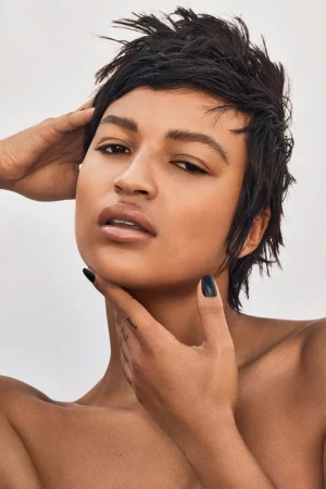 Close-up portrait of a person with short dark hair and light skin, hands near the neck, set against a pale background.
