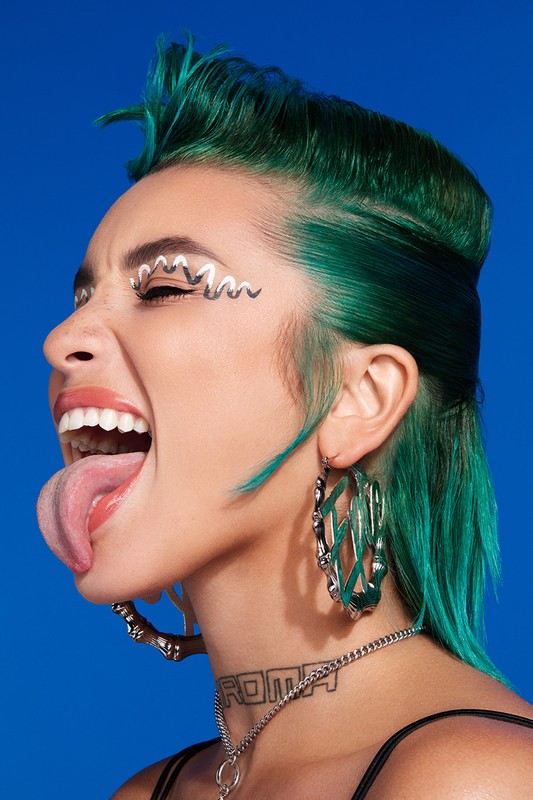 Profile portrait of a woman with vibrant green hair sticking her tongue out, wearing large hoop earrings against a solid blue background.