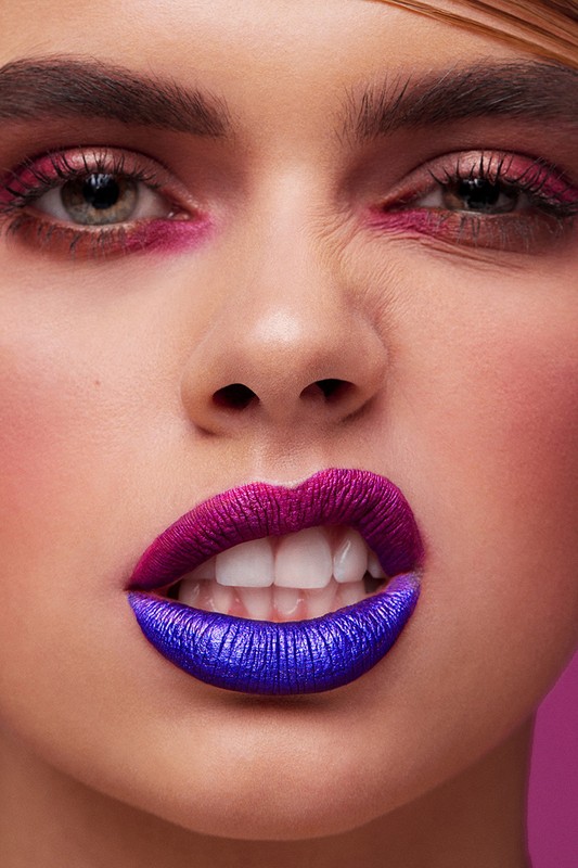 Close-up portrait of a woman with creative makeup. Her lips are painted with a duo-tone of purple and blue lipstick with pink eyeshadow.