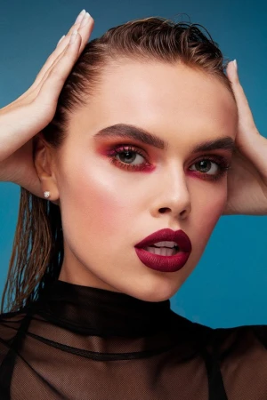 Close-up portrait of a fair-skinned adult woman with wet hair, bold eye and lip makeup against a blue backdrop.