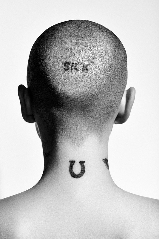 Back view of an adult with a shaved head featuring a tattoo of the word 'SICK,' with horseshoe and letter 'V' tattoos on their neck.