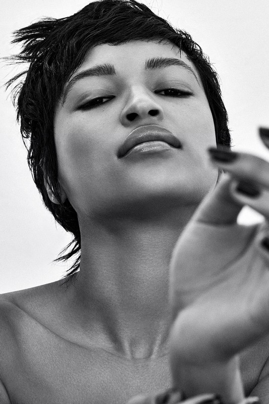 Striking black and white portrait of an adult female with a short, choppy hairstyle and a hand raised towards the camera in a simple studio.