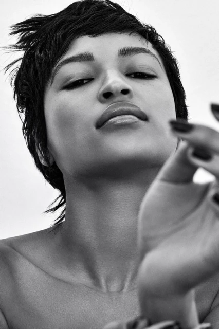 Striking black and white portrait of an adult female with a short, choppy hairstyle and a hand raised towards the camera in a simple studio.