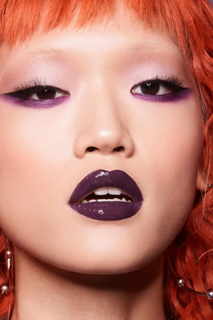 Close-up shot of an Asian woman with vibrant orange hair and dramatic purple makeup on eyes and glossy lips in a beauty context.