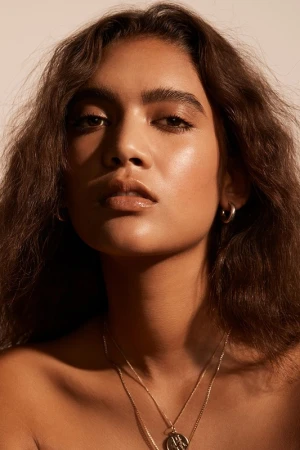 Close-up portrait of a woman with long wavy dark brown hair wearing layered gold necklaces against a warm neutral background.
