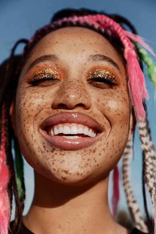 Close-up portrait of a smiling woman with freckles and colorful braids against a clear blue sky, highlighting her radiant complexion and festive makeup.
