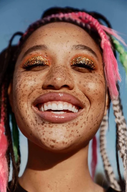 Close-up portrait of a smiling woman with freckles and colorful braids against a clear blue sky, highlighting her radiant complexion and festive makeup.