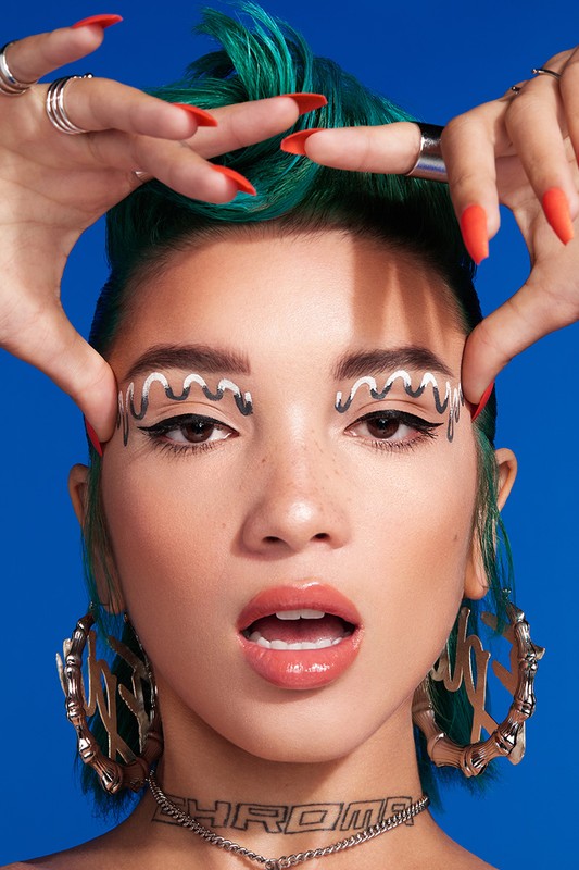 Close-up portrait of a woman with teal hair, makeup with silver accents, large hoop earrings, and a 'CHROME' necklace, against a blue backdrop.