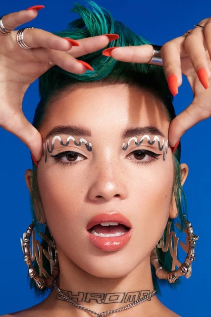 Close-up portrait of a woman with teal hair, makeup with silver accents, large hoop earrings, and a 'CHROME' necklace, against a blue backdrop.