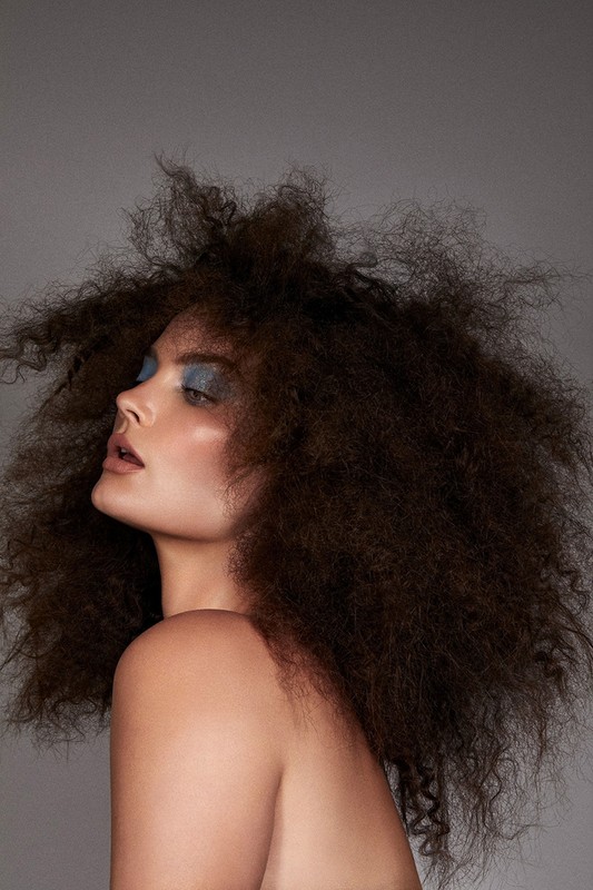 Portrait of a woman with voluminous dark curly hair and blue eyeshadow against a plain grey background by photographer Brooke Nipar.