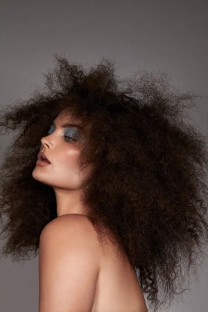 Portrait of a woman with voluminous dark curly hair and blue eyeshadow against a plain grey background by photographer Brooke Nipar.