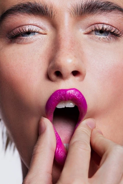Close-up of a fair-skinned adult female with pink lipstick, eye makeup, and fingers pulling down her lower lip in a beauty shot.