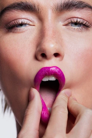 Close-up of a fair-skinned adult female with pink lipstick, eye makeup, and fingers pulling down her lower lip in a beauty shot.