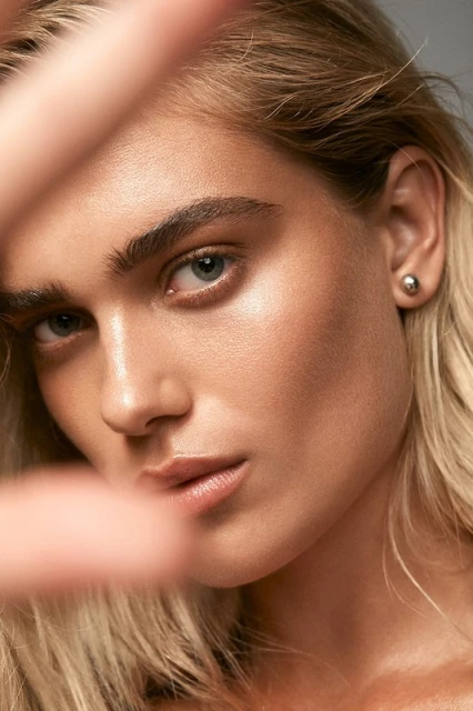 Close-up studio portrait of a young adult female model with blonde hair, wearing earrings, posing with her hand partially covering her face.