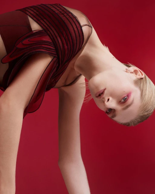Fashion portrait of a fair-skinned blonde woman in a burgundy garment, leaning forward against a solid red backdrop in a photo studio.