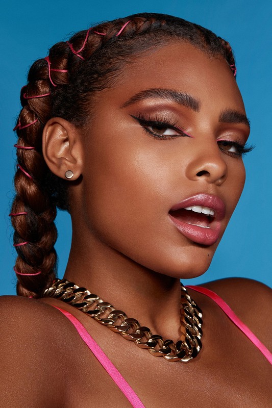 Close-up portrait of a woman with braided hair, pink accents, winged eyeliner, gold chain necklace, and blue background.