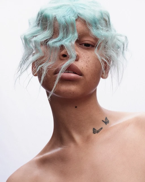 Portrait of a freckled woman with light blue wavy hair and butterfly tattoos on her neck against a white backdrop in a photo studio.