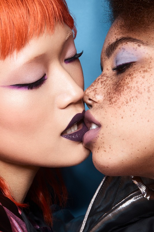 Close-up of two women with creative makeup, one with red hair and the other with short dark hair, against a blue background.