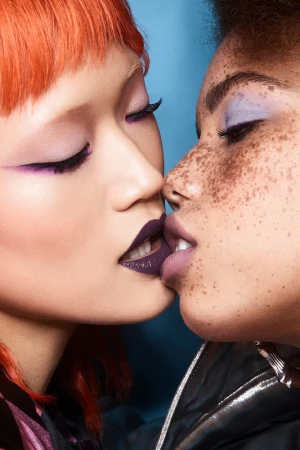 Close-up of two women with creative makeup, one with red hair and the other with short dark hair, against a blue background.