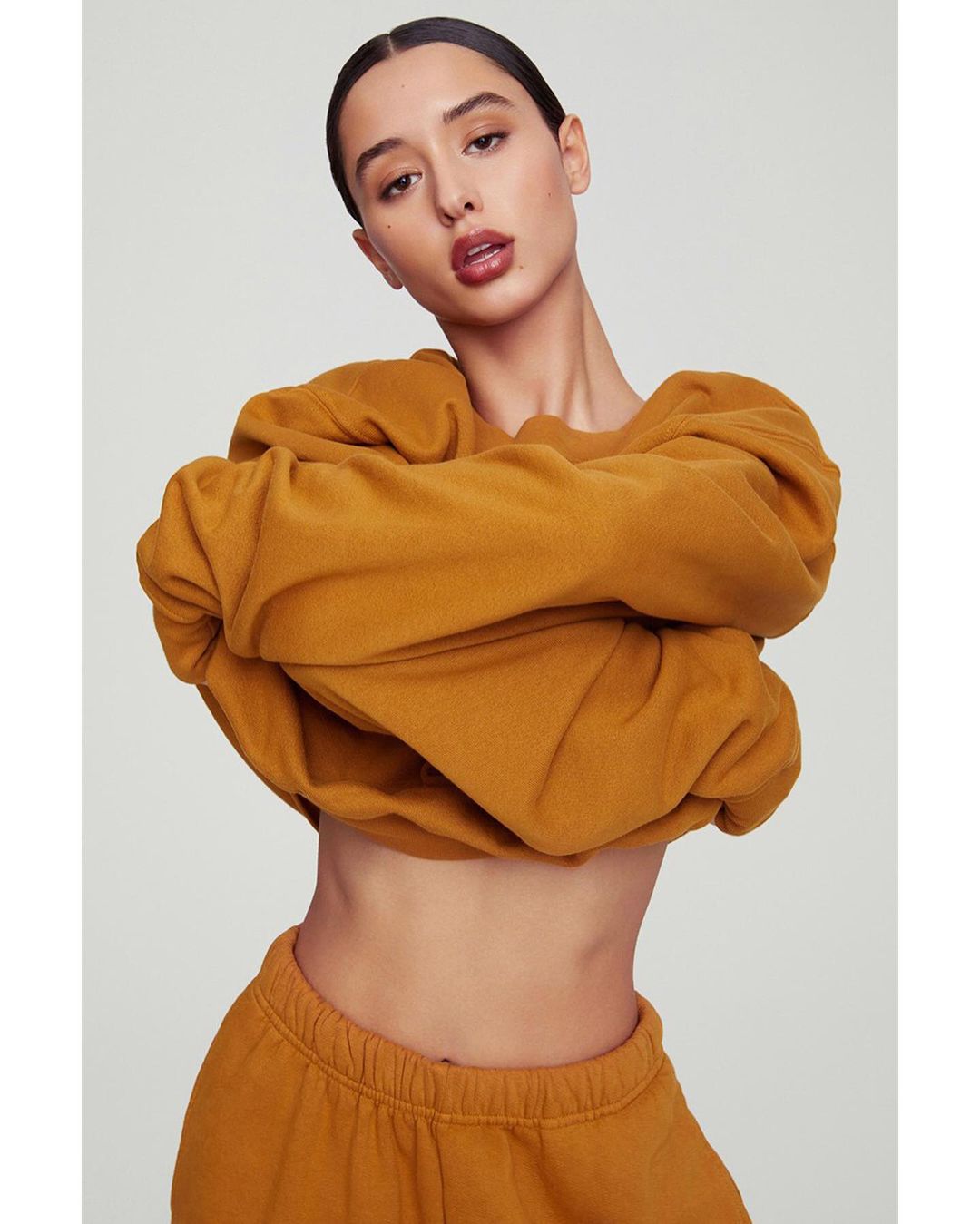 Woman in an orange cropped sweatshirt and sweatpants poses against a neutral background. Close-up studio shot. Fashion shoot.