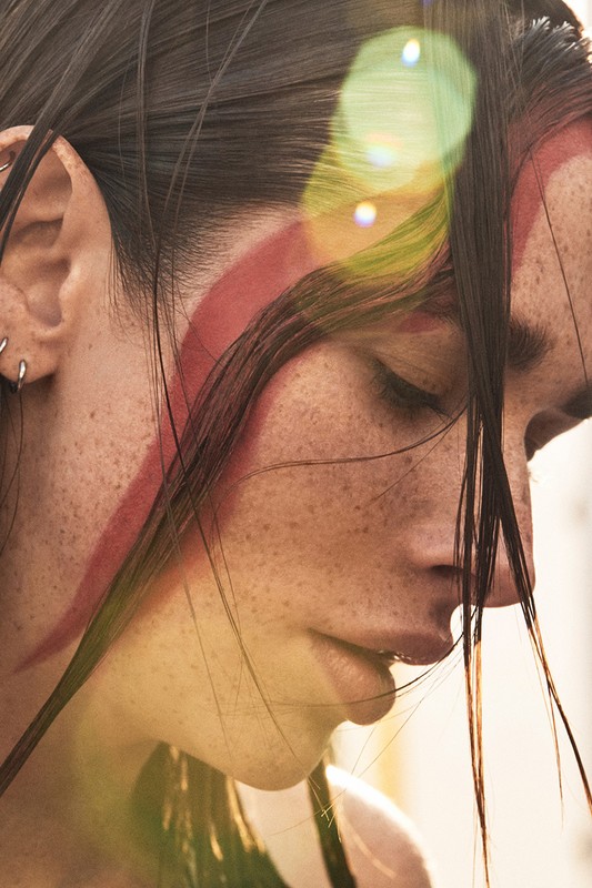 Profile view of a woman with freckled skin, dark hair, and red paint on her face, bathed in soft, natural light with a warm lens flare.