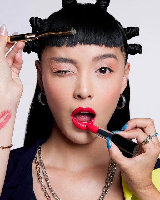 Cosmetic advertisement featuring a young adult woman with bold makeup and styled hair posing in a beauty studio with props.