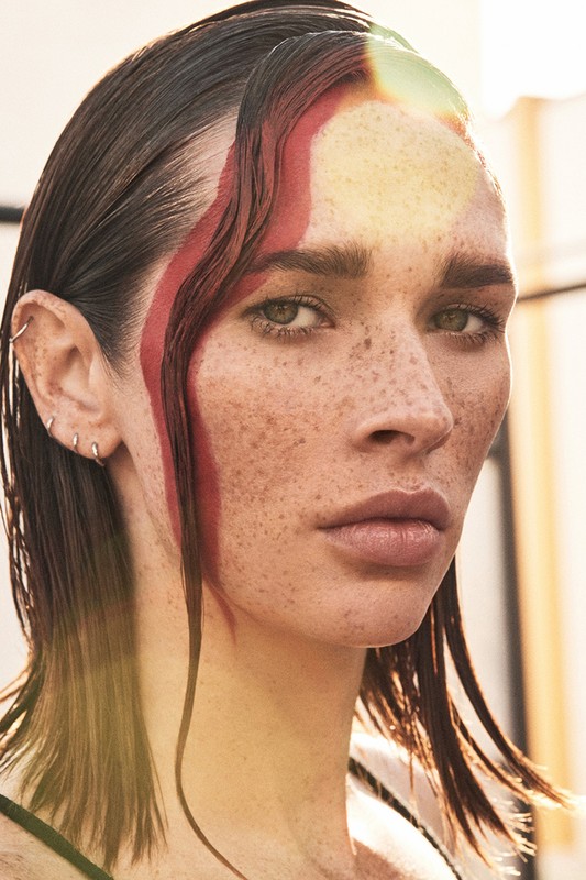 Close-up portrait of an adult woman with freckles, sleek wet hair, and a bold red streak of paint on her forehead, captured outdoors.