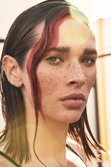 Close-up portrait of an adult woman with freckles, sleek wet hair, and a bold red streak of paint on her forehead, captured outdoors.